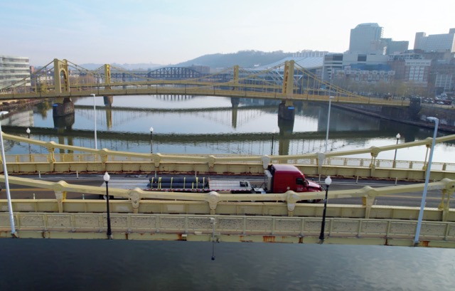 Birds-eye-view photo of red JLE truck hauling flatbed load across a bridge towards a big city