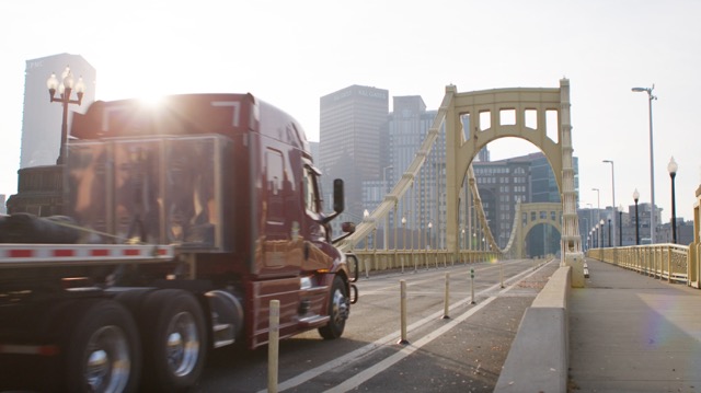 Action photo of red JLE truck hauling flatbed load across a bridge towards a big city